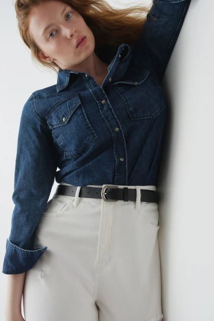 A fashion shot of a young adult woman with red hair wearing a denim shirt and white jeans indoors against a white wall.