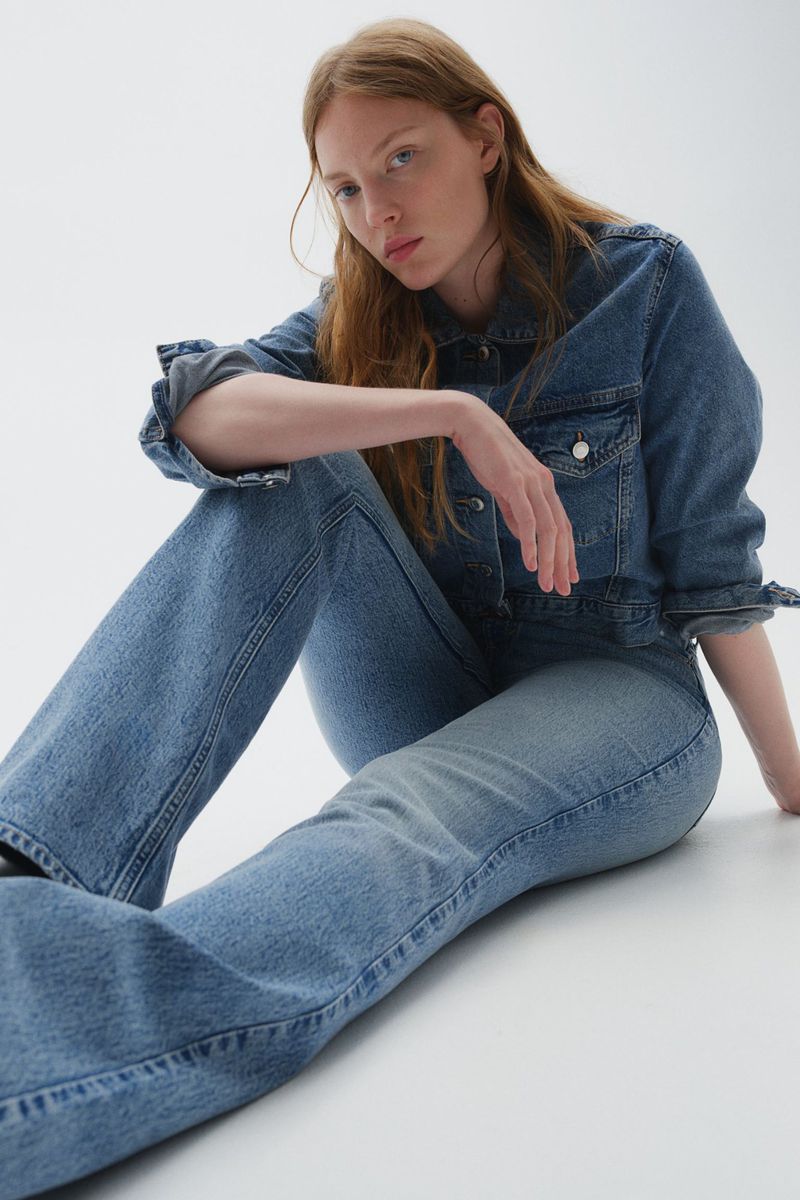 Model seated on a white studio floor wearing a matching blue denim jacket and flared denim jeans.