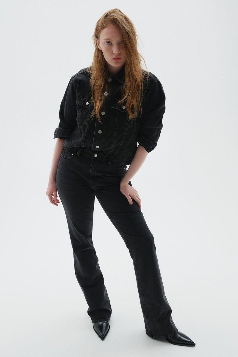 A full-length shot of a woman with red hair, wearing a black denim jacket and jeans, posing against a white backdrop in a fashion editorial style.