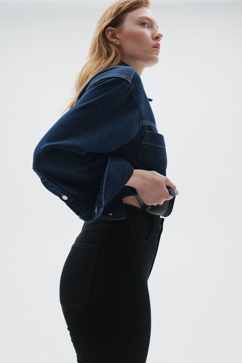 A slender woman with long red hair poses sideways in a denim jacket and black jeans against a bright white backdrop in a fashion shot.