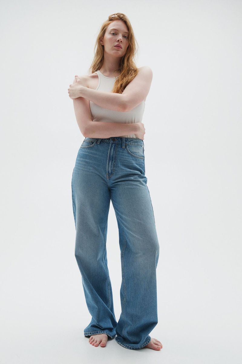 Model Anneta Popova wearing a light tank top and wide-leg blue jeans standing barefoot against a neutral studio backdrop.
