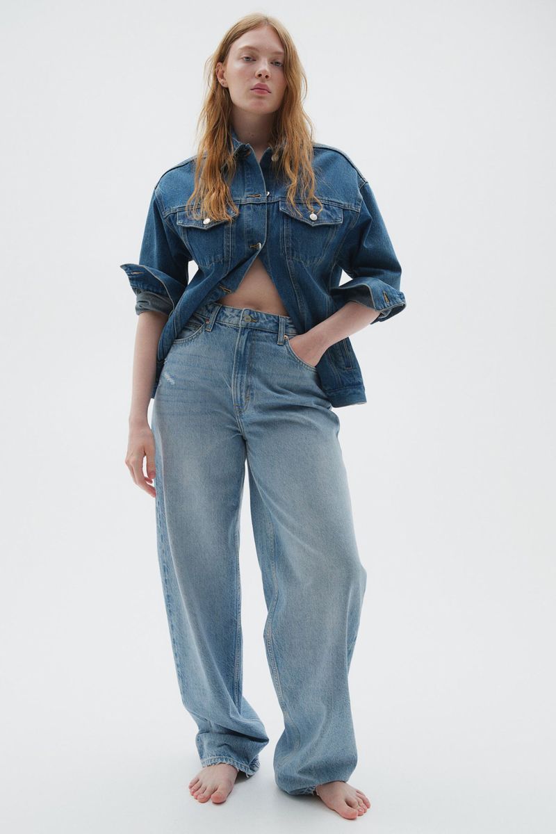Woman in light-wash jeans and a dark denim jacket stands barefoot on a white background in a fashion shot.