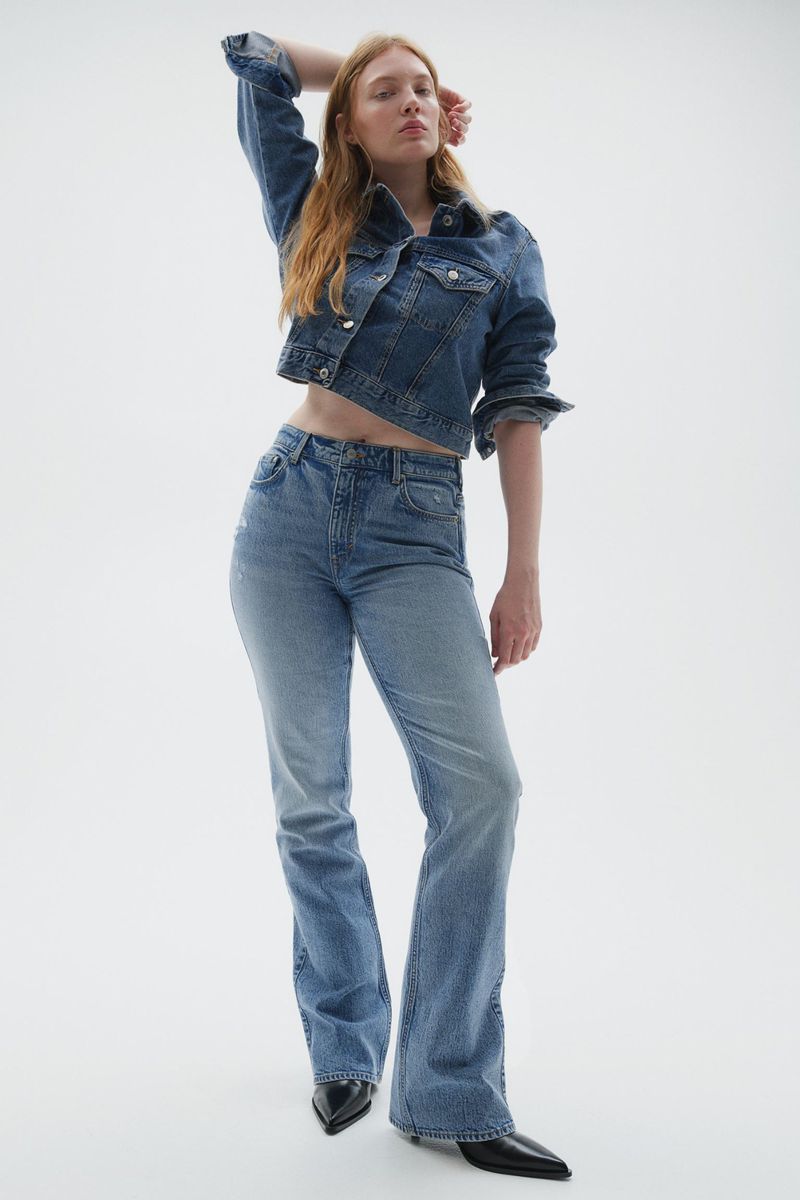 A redhead woman posing in a jean jacket and jeans with a white backdrop, showcasing a modern fashion ensemble with neutral tones.