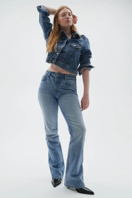 A redhead woman posing in a jean jacket and jeans with a white backdrop, showcasing a modern fashion ensemble with neutral tones.