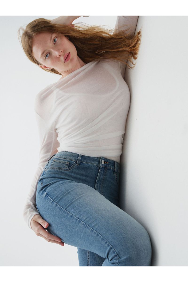 Fashion shot of a red-haired woman in a sheer top and blue jeans, posing against a white wall with her hand on her hip.