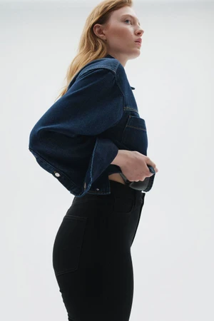 Side profile of a red-haired woman wearing a dark blue cropped denim jacket and high-waisted black jeans against a white background.