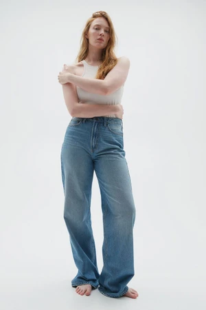 Model Anneta Popova wearing a light tank top and wide-leg blue jeans standing barefoot against a neutral studio backdrop.