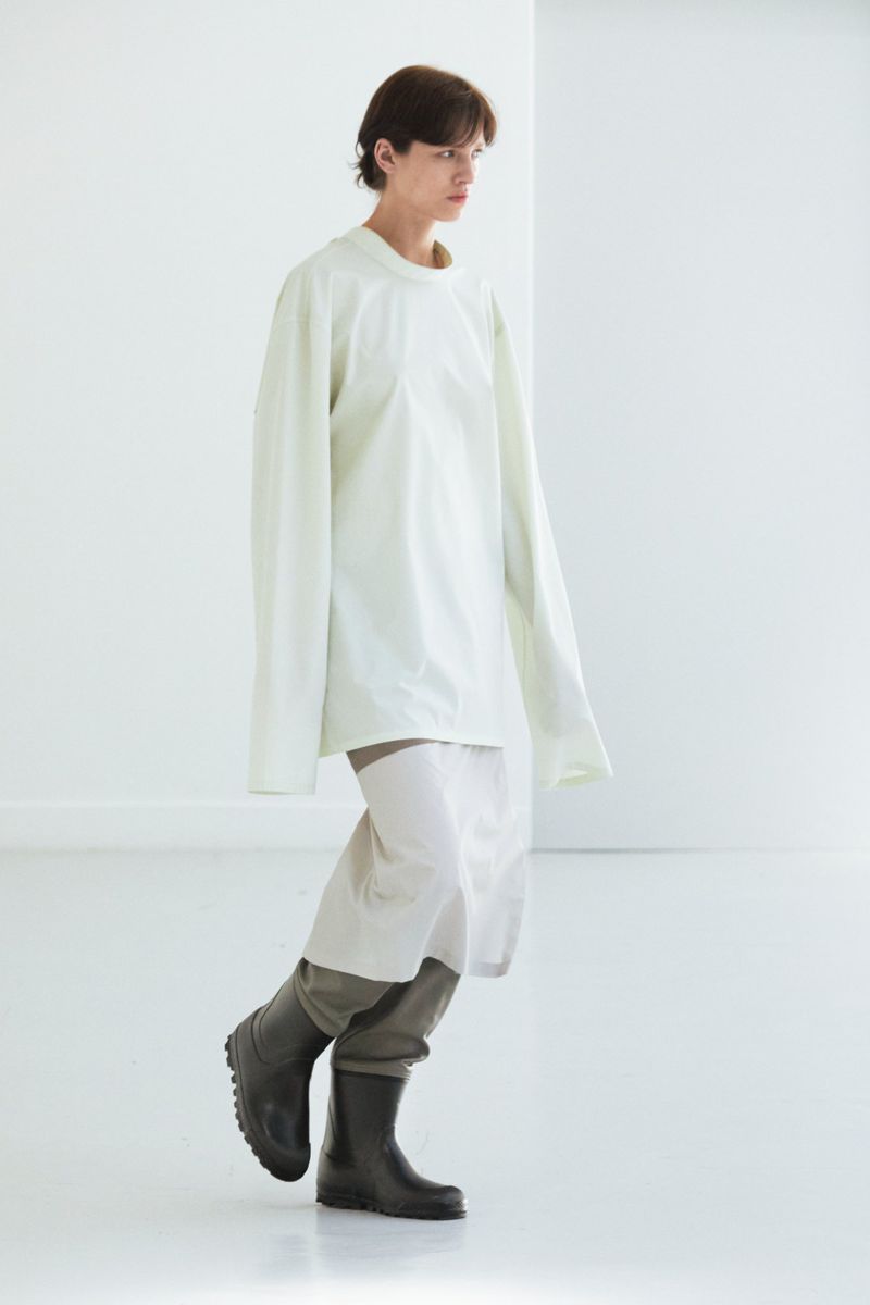A person with dark hair is walking in a bright white space, wearing a long-sleeved white shirt, light pants, and black boots.