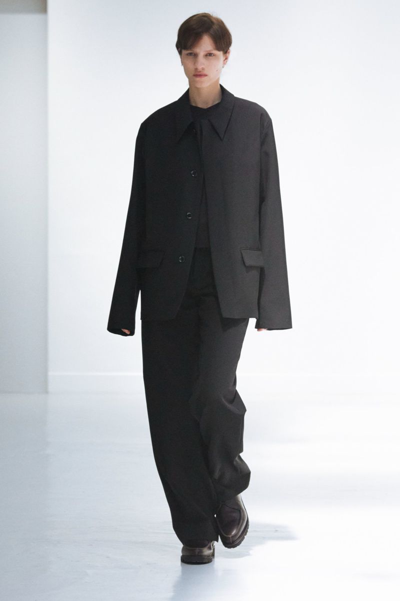 Runway fashion show featuring a slender adult woman wearing a black tailored suit with a buttoned shirt and dress shoes.