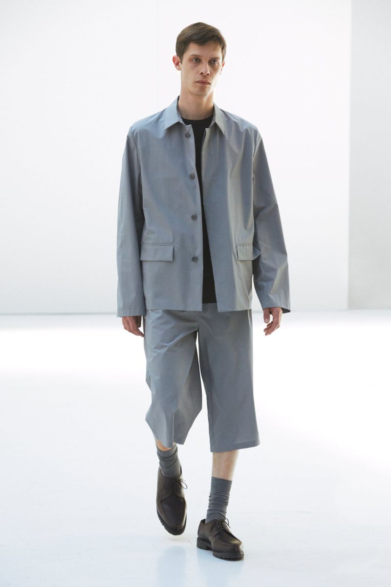 Male fashion model showcases a light grey jacket and shorts outfit, paired with socks and dress shoes, against a minimalist white backdrop.