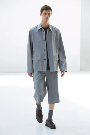 Male fashion model showcases a light grey jacket and shorts outfit, paired with socks and dress shoes, against a minimalist white backdrop.