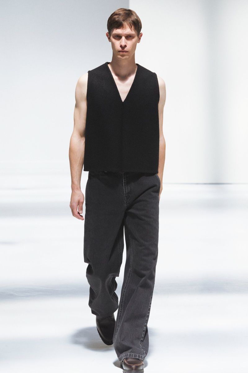 Male fashion model walks a runway in a black vest and gray jeans. Full-length shot with bright white seamless background.