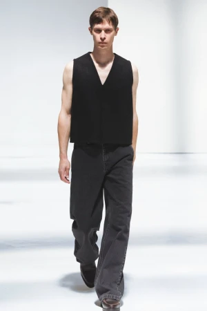 Male fashion model walks a runway in a black vest and gray jeans. Full-length shot with bright white seamless background.