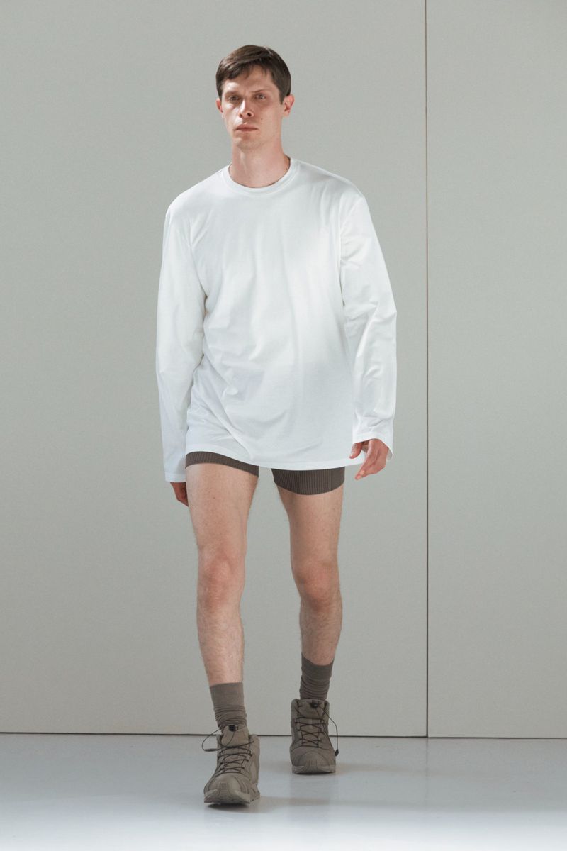 Male fashion model wearing a white long-sleeve shirt, brown shorts, socks, and shoes poses in a brightly lit white studio.