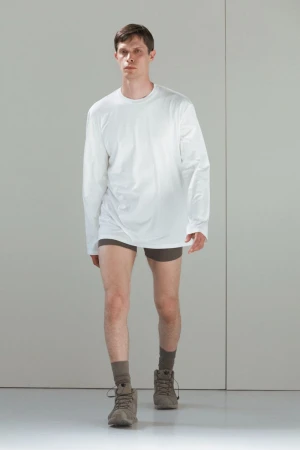 Male fashion model wearing a white long-sleeve shirt, brown shorts, socks, and shoes poses in a brightly lit white studio.