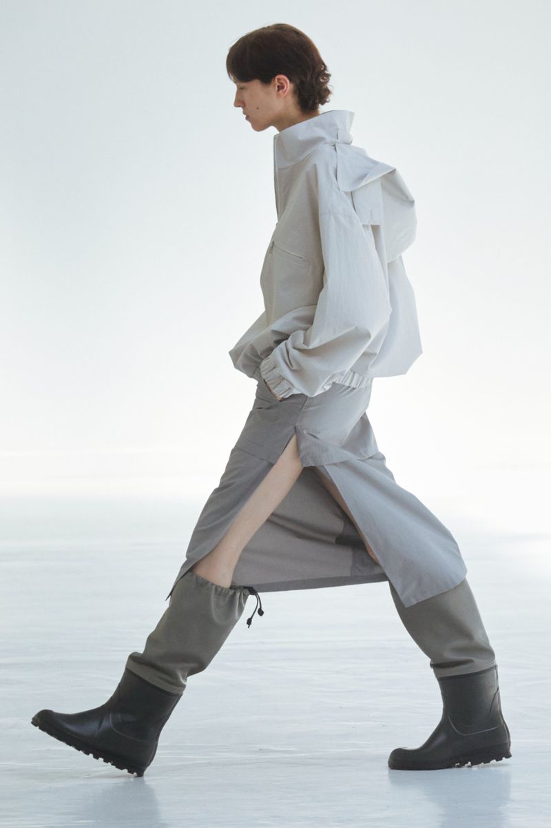 Fashionable adult strides forward in a light grey jacket, grey midi skirt with a side slit, and black boots against a light background.