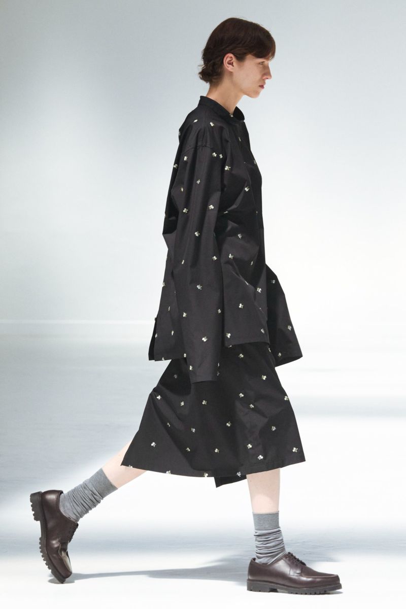 A woman in a black patterned shirt and skirt walks in front of a white background in a fashion runway or studio setting, wearing brown shoes.