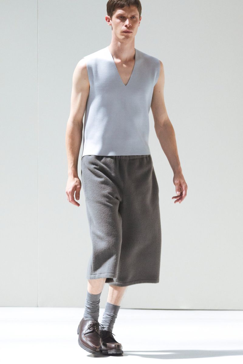 Male fashion model showcases a light blue sleeveless top and gray culottes in a clean studio setting, complemented by brown leather shoes.