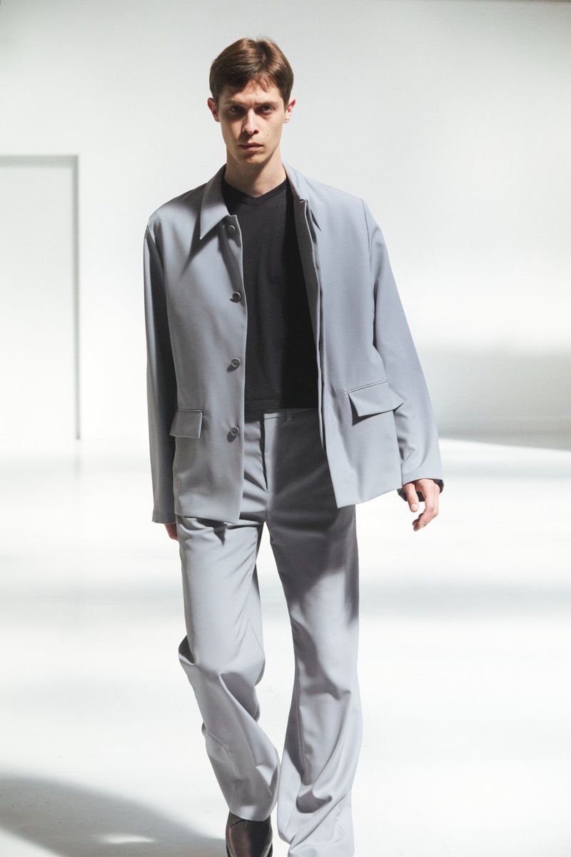 Male model walking in a light grey tailored jacket, matching trousers, and a black shirt in a bright white studio space.