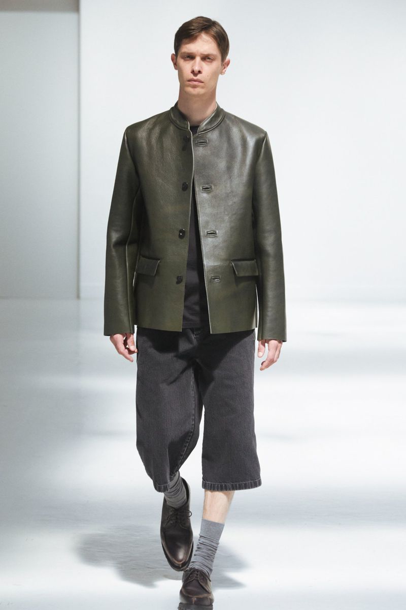 A slender adult male presents fashion, wearing an olive leather jacket, dark culottes, grey socks, and dress shoes in a studio setting.