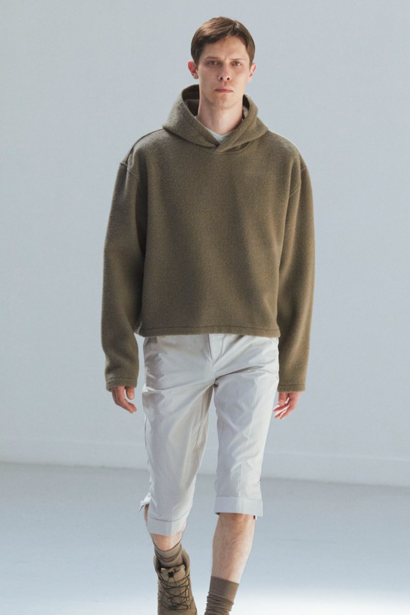 Male model in a studio wearing a cropped olive green hoodie, light tan capri pants, and brown boots, posing in a full shot.