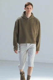 A male model walking forward in an olive fleece hoodie and light grey cropped pants in a bright studio.