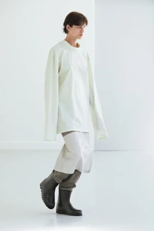 Model wearing an oversized white top and dark rubber boots against a plain white studio background.