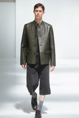 Male model in an olive green leather jacket and cropped dark trousers walking against a white studio background.