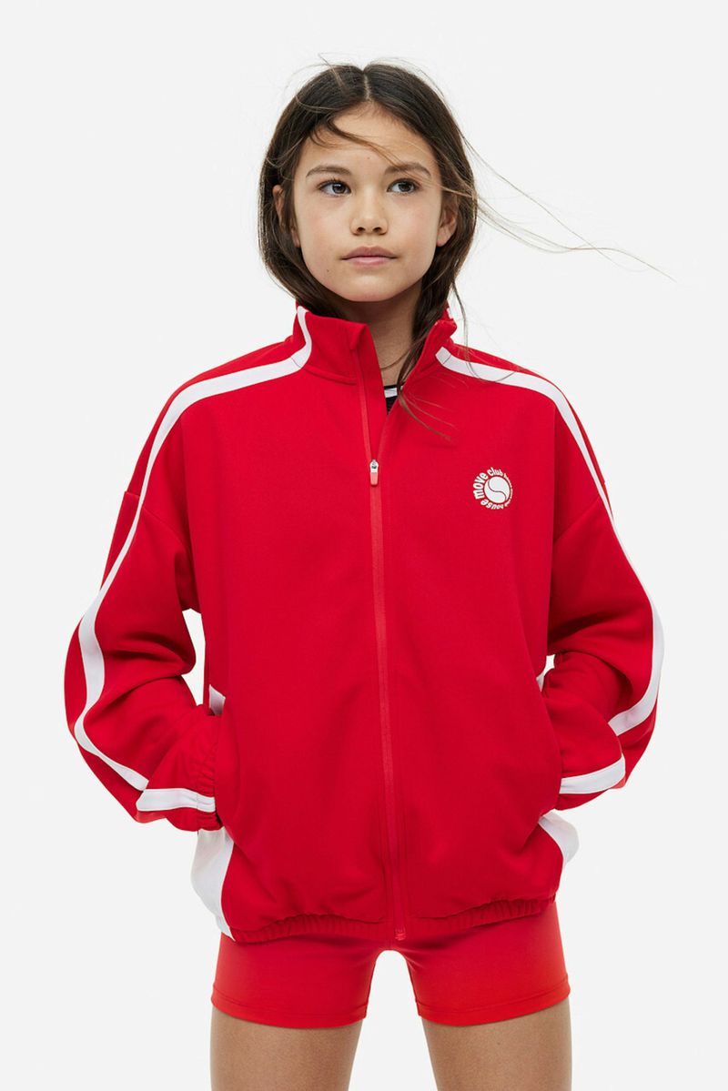Young girl in a red track jacket with white stripes and red shorts posing against a white background in a studio setting.