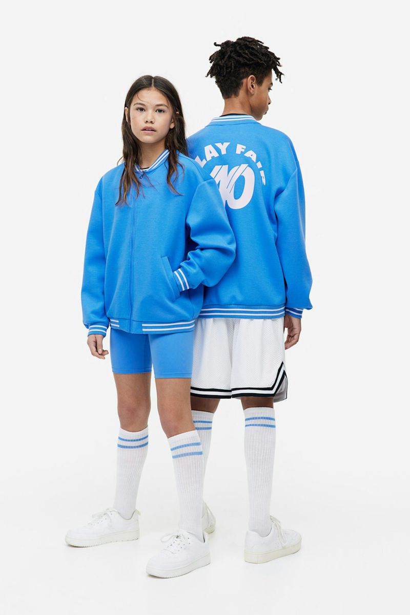 Two young fashion models in matching letterman jackets, shorts, socks, and sneakers pose on a seamless white background.