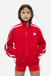 Young girl wearing a bright red zip-up track jacket with white sleeve stripes and matching red shorts against a studio backdrop.