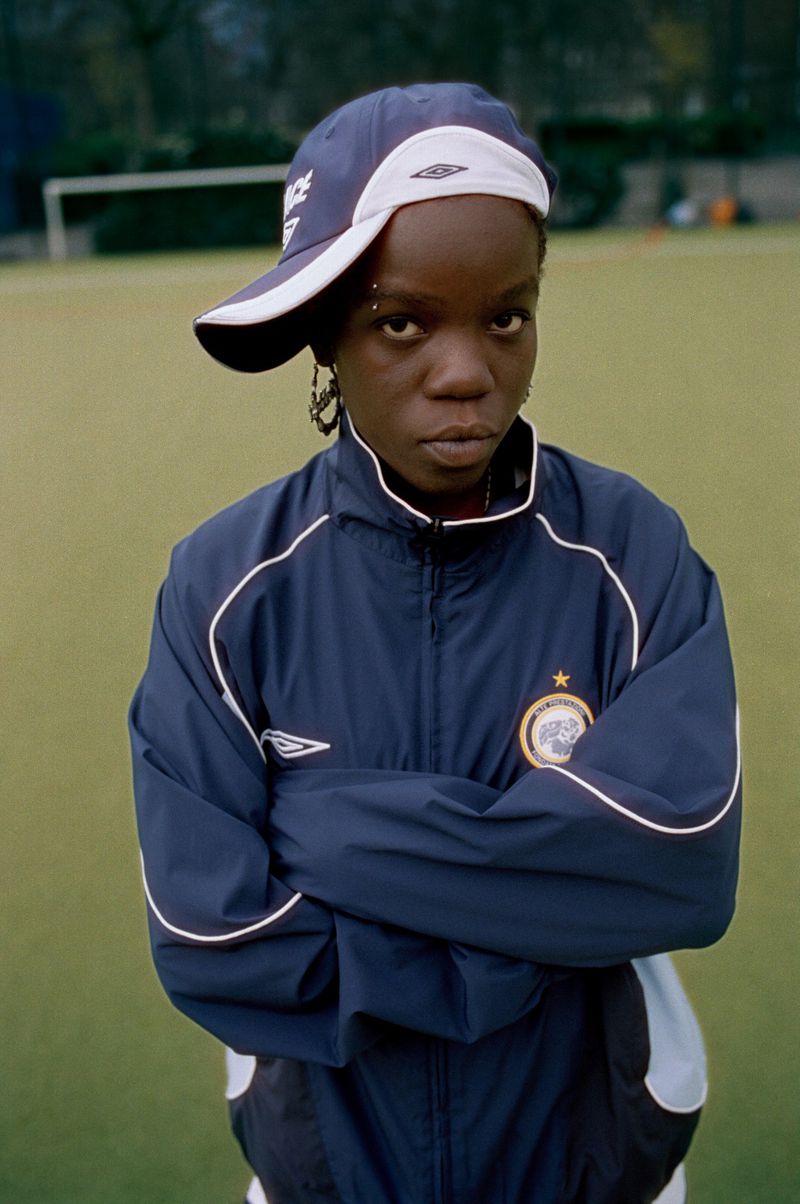 Portrait of a young Black woman wearing a sports cap and jacket, standing with arms crossed against a green lawn backdrop.