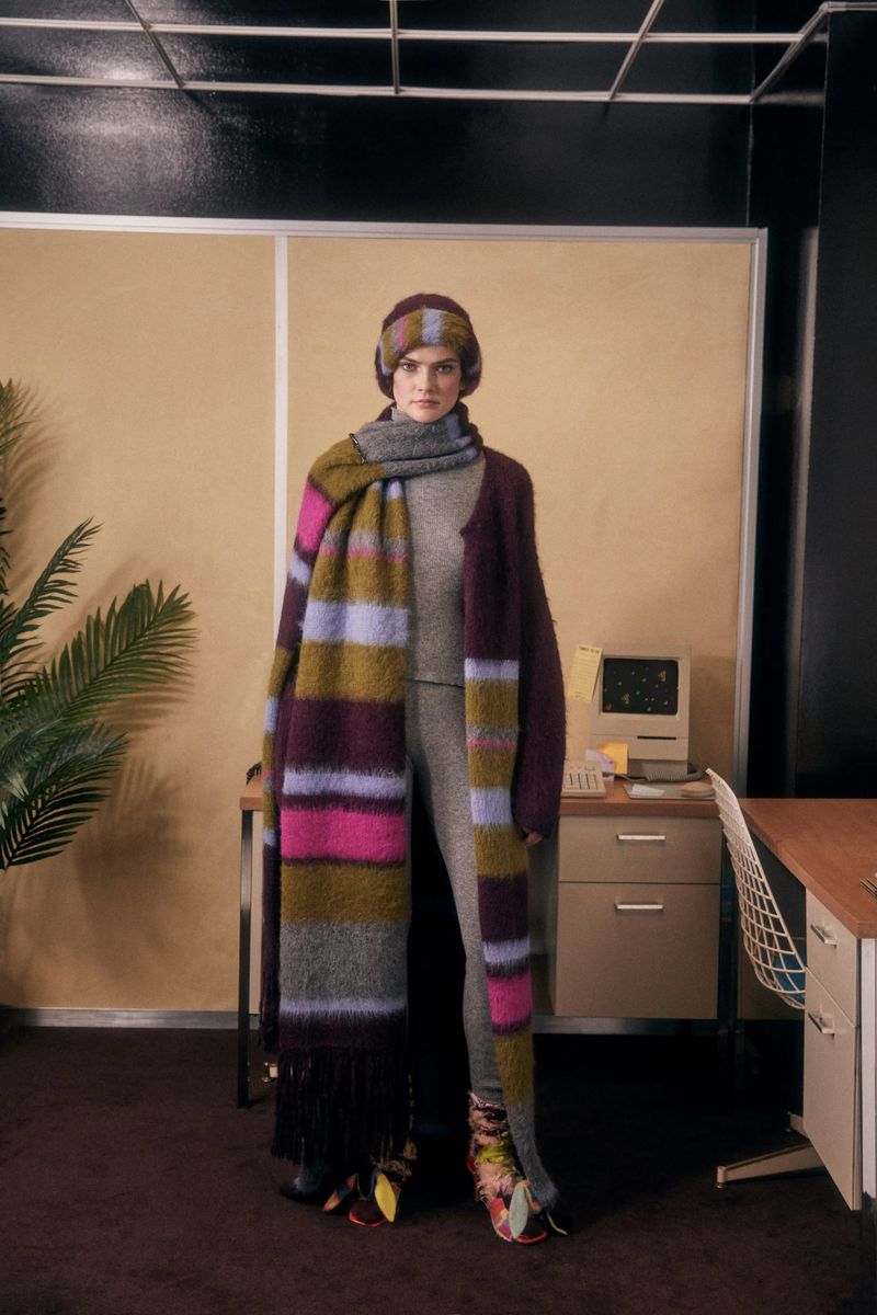 Fashionable woman in indoor setting wears a grey knit outfit, a colorful striped scarf, and a matching hat, posing in an office.