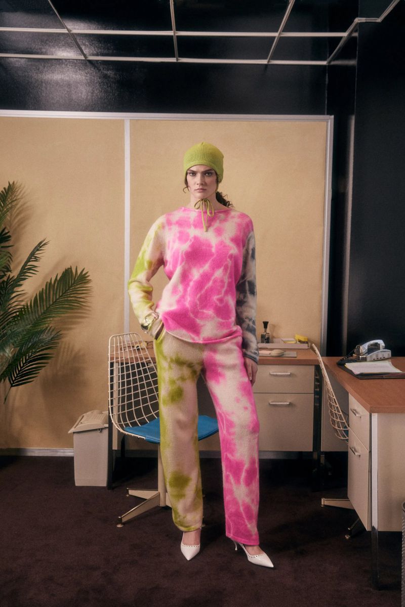 A woman models colorful tie-dye knitwear with white heels, posing in a room with a retro desk, chair, and indoor plant.