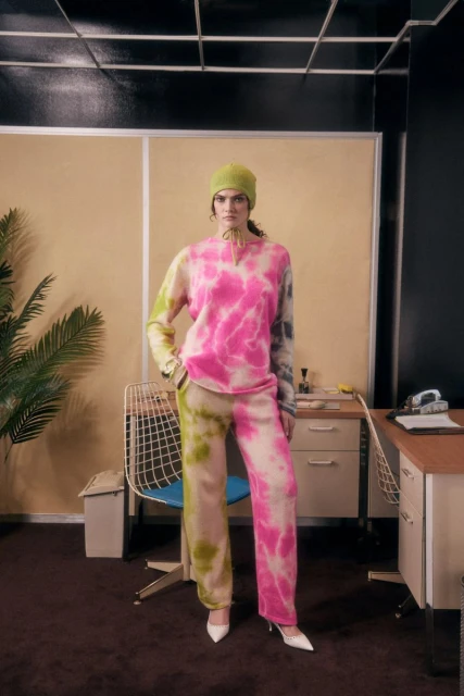 A woman models colorful tie-dye knitwear with white heels, posing in a room with a retro desk, chair, and indoor plant.