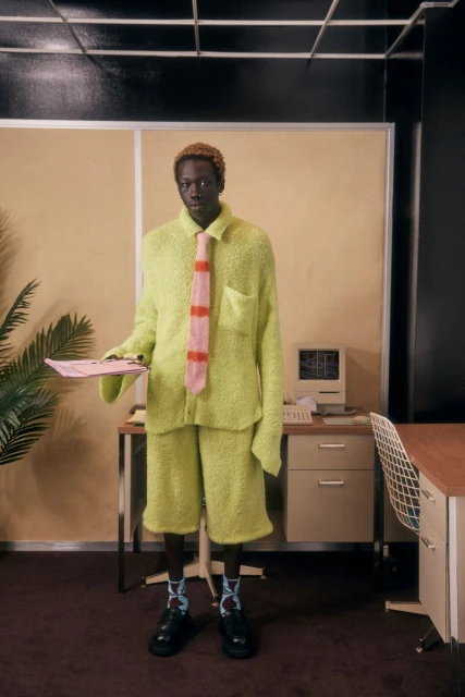 Man in bright green shorts suit, tie, and black shoes stands in office holding pink papers, with retro computer and office furniture.
