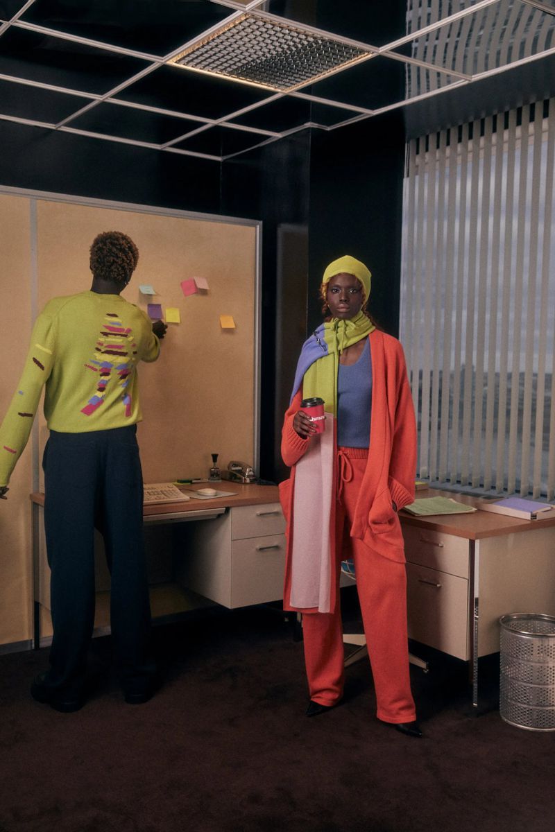Two stylish adults in an office. An adult female wearing a yellow hat and orange suit stands next to a desk while holding a coffee cup.