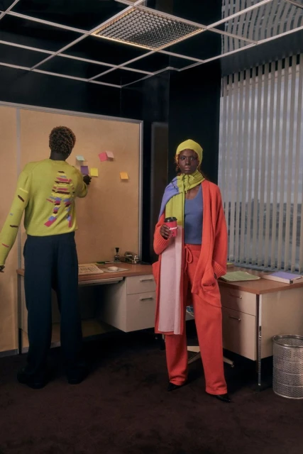 Two stylish adults in an office. An adult female wearing a yellow hat and orange suit stands next to a desk while holding a coffee cup.