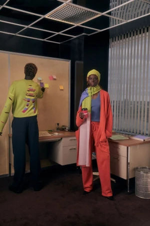 Two stylish adults in an office. An adult female wearing a yellow hat and orange suit stands next to a desk while holding a coffee cup.
