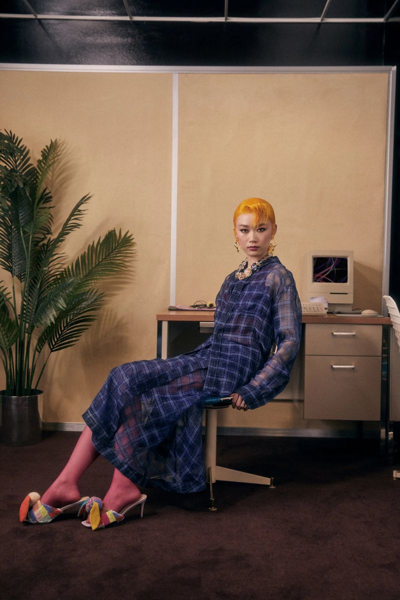 Woman with orange hair sits sideways in a blue-plaid dress, wearing colorful high heels; an office setting with a computer and a potted plant.