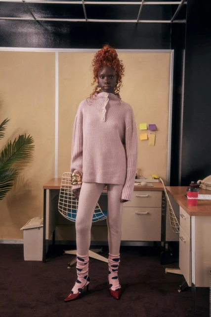 A redhead female model is posed in front of a retro office setup, wearing a light pink ribbed sweater, leggings, patterned socks, and red heels.