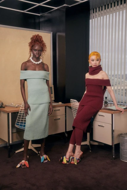 Two fashion models pose in knit dresses and statement shoes in an office-inspired setting with desks and a neutral color palette.