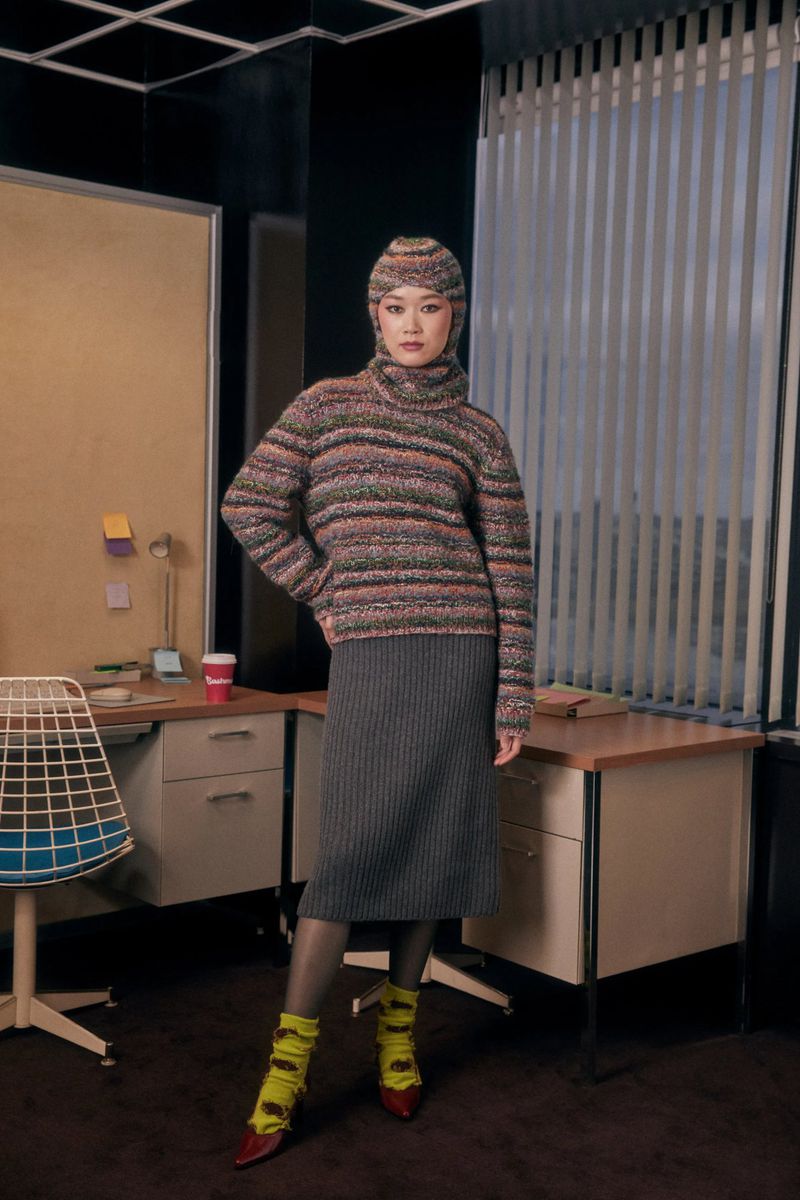 Adult female poses in a knit hat, sweater, and skirt near a desk. She wears yellow socks with red shoes in an indoor office environment.