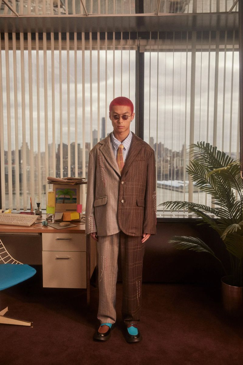 Adult male with dyed red hair stands wearing a tan tie, glasses, and a brown and tan suit in front of an office desk and window.