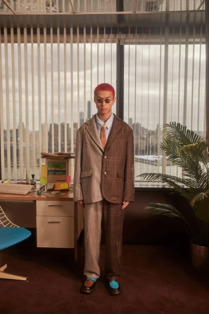 Adult male with dyed red hair stands wearing a tan tie, glasses, and a brown and tan suit in front of an office desk and window.