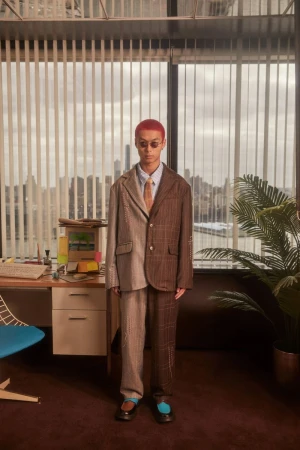 Adult male with dyed red hair stands wearing a tan tie, glasses, and a brown and tan suit in front of an office desk and window.