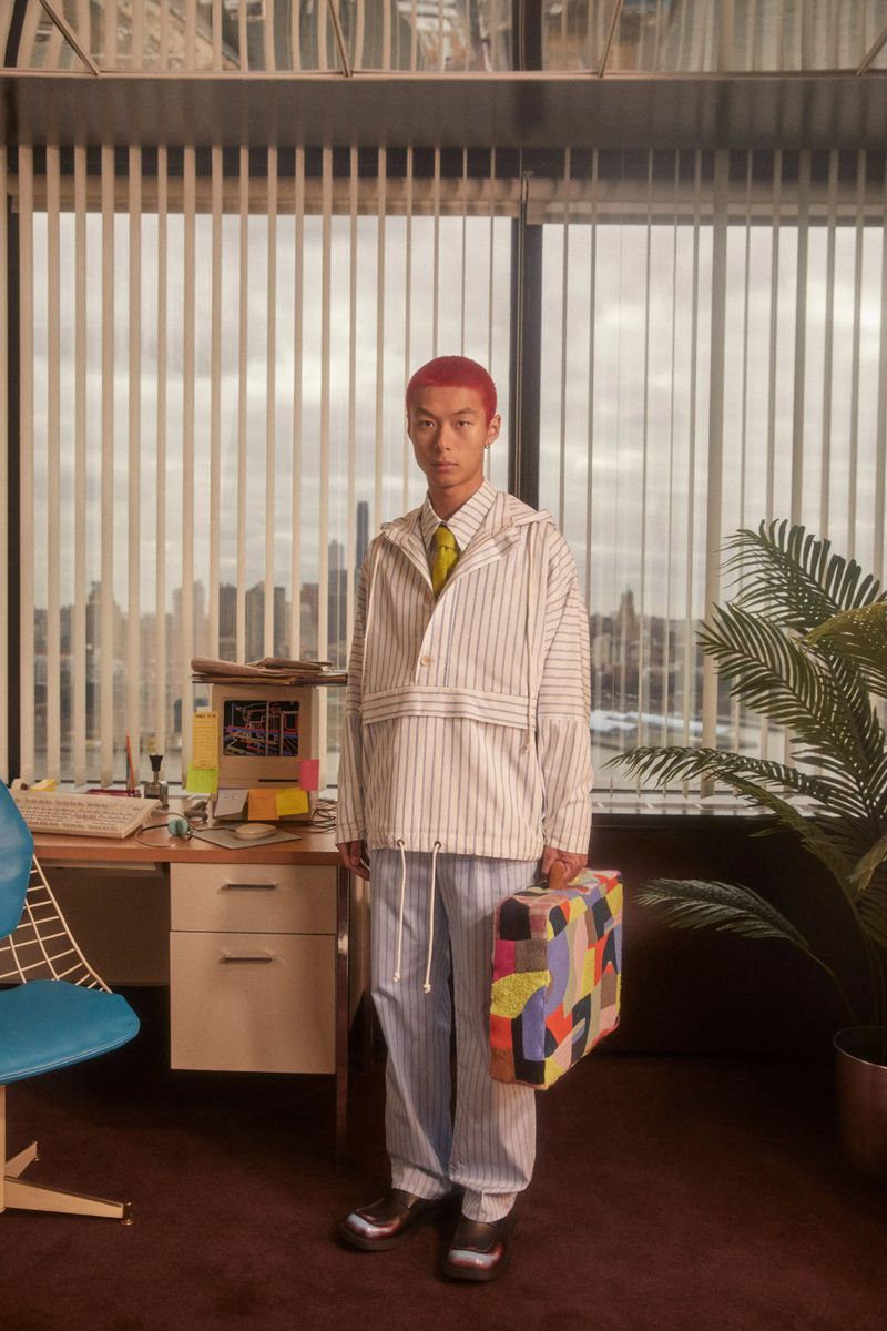 A person with red hair stands in an office setting holding a colorful briefcase, wearing a striped jacket and pants with a neutral tie.