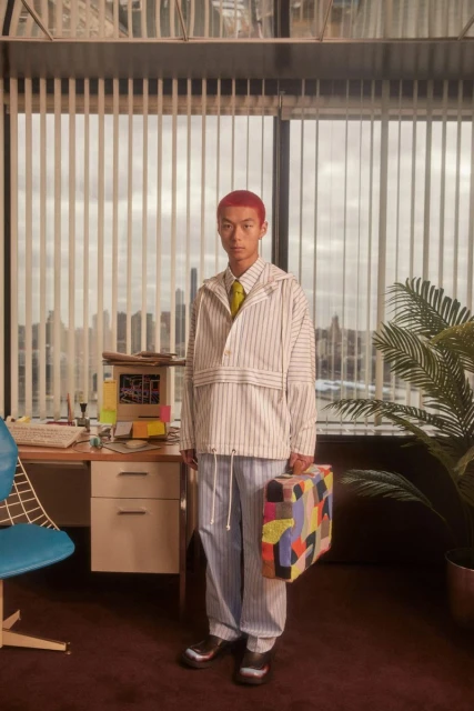 A person with red hair stands in an office setting holding a colorful briefcase, wearing a striped jacket and pants with a neutral tie.