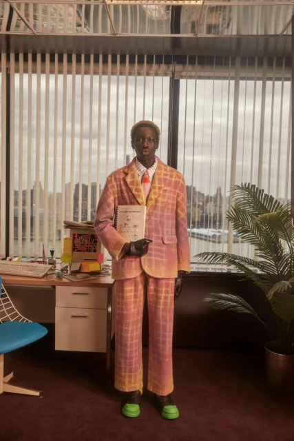 A dark-skinned male stands in a windowed office wearing a pink and yellow plaid suit and holding a book while facing forward.