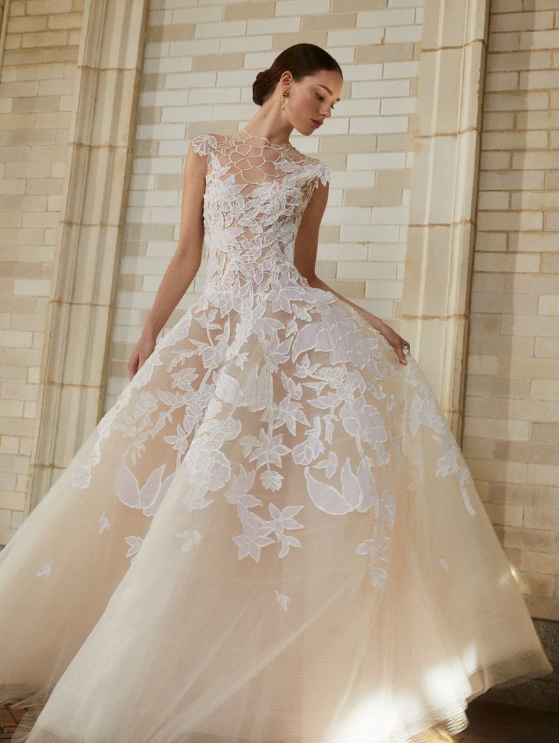 Elegant woman in a light peach ballgown with lace floral details, posing in an indoor setting with natural light.